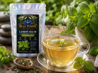 A Blue Temple Botany Lemon Balm Herbal Tea pouch next to a glass cup of brewed lemon balm tea on a wooden surface with fresh mint leaves and loose tea.