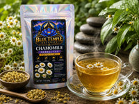 Blue Temple Botanicals Chamomile Herbal Tea pouch shown next to a glass cup of brewed chamomile tea with chamomile flowers floating on the surface, surrounded by loose chamomile seeds, a wooden spoon, and fresh chamomile flowers in a garden setting.
